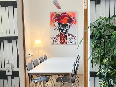 Blick durch einen Türrahmen in einen Raum mit großem Arbeitstisch mit Stühlen, gemütlicher Beleuchtung und dem namensgebenden modernen Bild an der Wand, das die Rückansicht einer Schwarzwälderin mit Bollenhut zeigt.