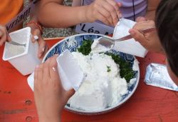 Ein Tisch auf dem ein Teller steht. Der Teller wird durch drei Kinder mit Quark befüllt.