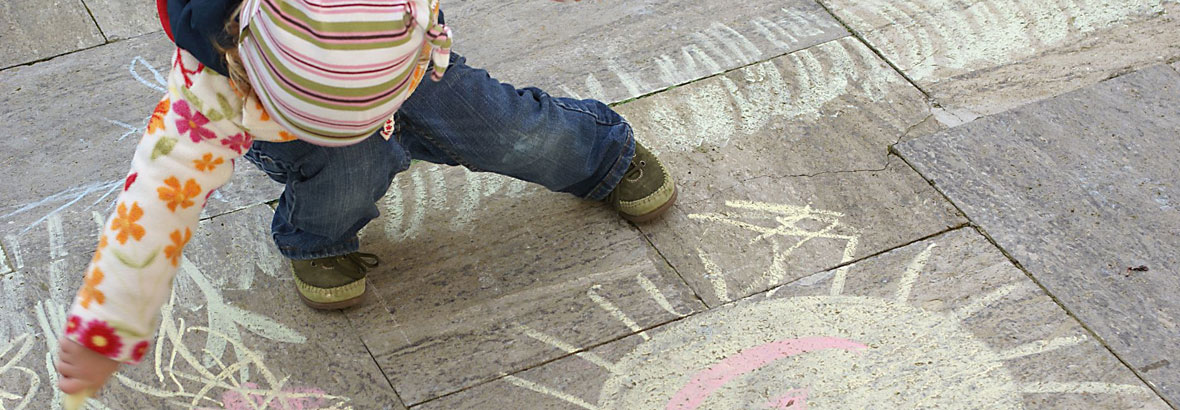 Ein Kleinkind malt mit Strassenmalkreide eine Sonne auf den Boden.